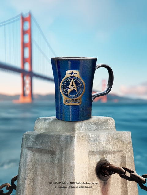 A handcrafted blue stoneware mug with the Starfleet Command logo from the Star Trek franchise, displayed on a textured platform with a bridge in the background.